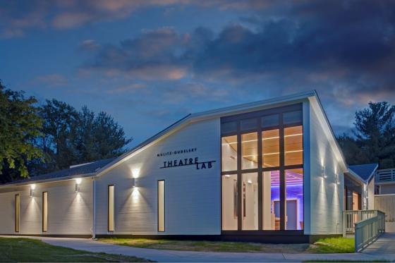 The outside of the Mulitz-Gudelsky Theatre Lab, a white, modern, minimalist building, lit up on a cloudy evening.